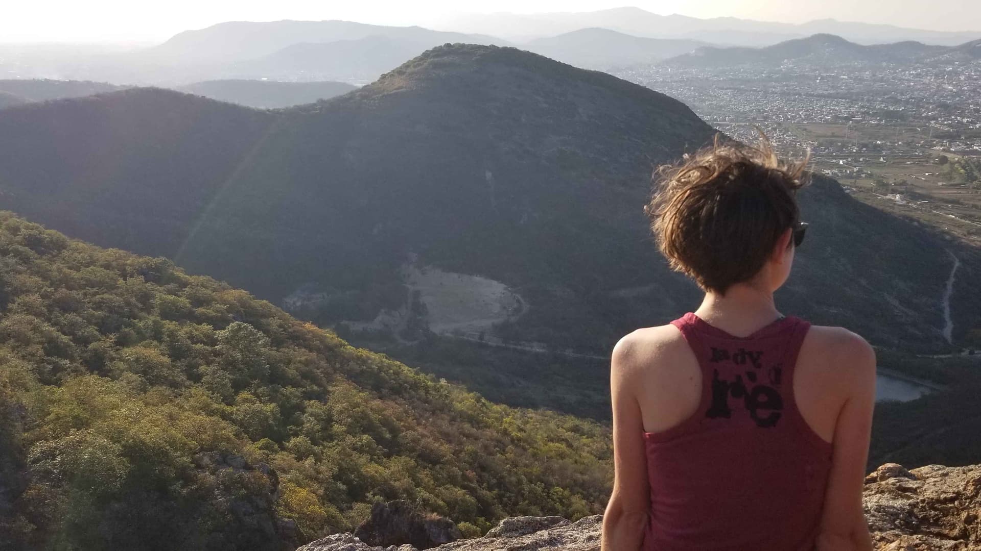 A person with short hair and wearing a red tank top sits facing away from the camera. In the distance are shrub covered hills and a city landscape.