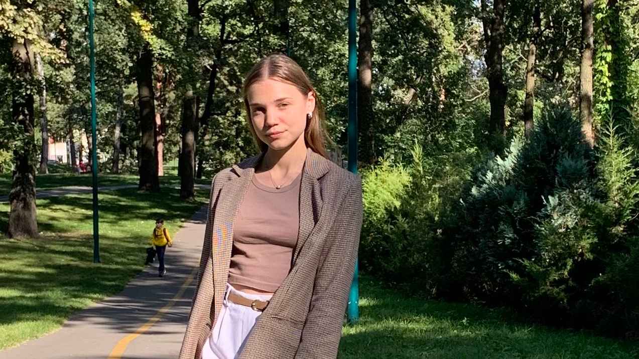 Valeriia Ivanova stands on a pathway with lush green grass and trees behind her.