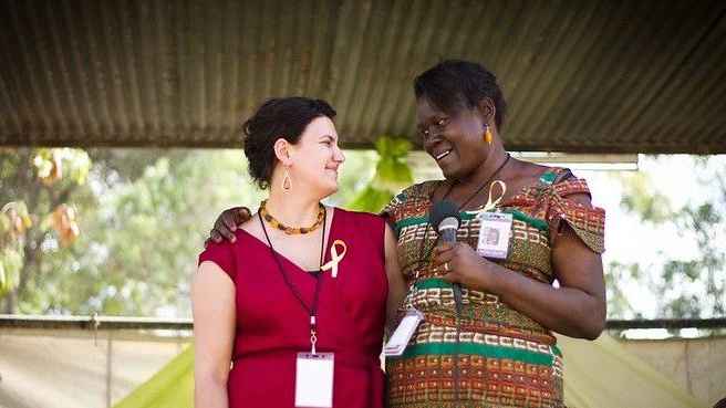 Photo of Jensine and Leah in colorful patterned clothes standing in Kenya under trees.  Leah holds microphone and they are looking into each others eyes with deep friendship and connection 
