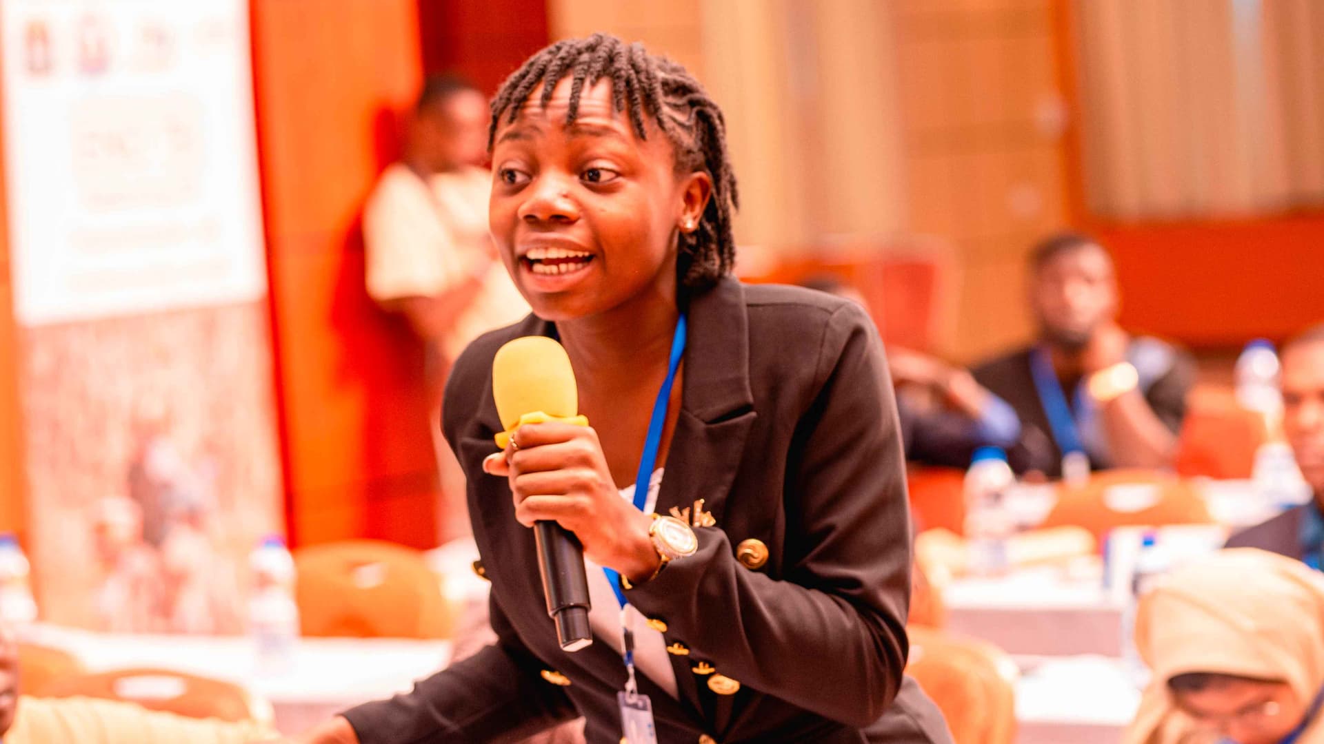 Liz, a woman with short dark hair in a black blazer, smiles and speaks before a conference audience. She is holding a mic.